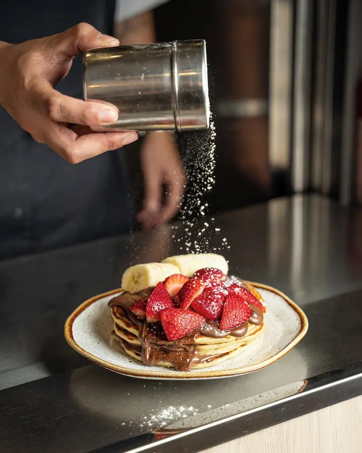 Hand dusting sugar over pancakes with Nutella, strawberries and banana