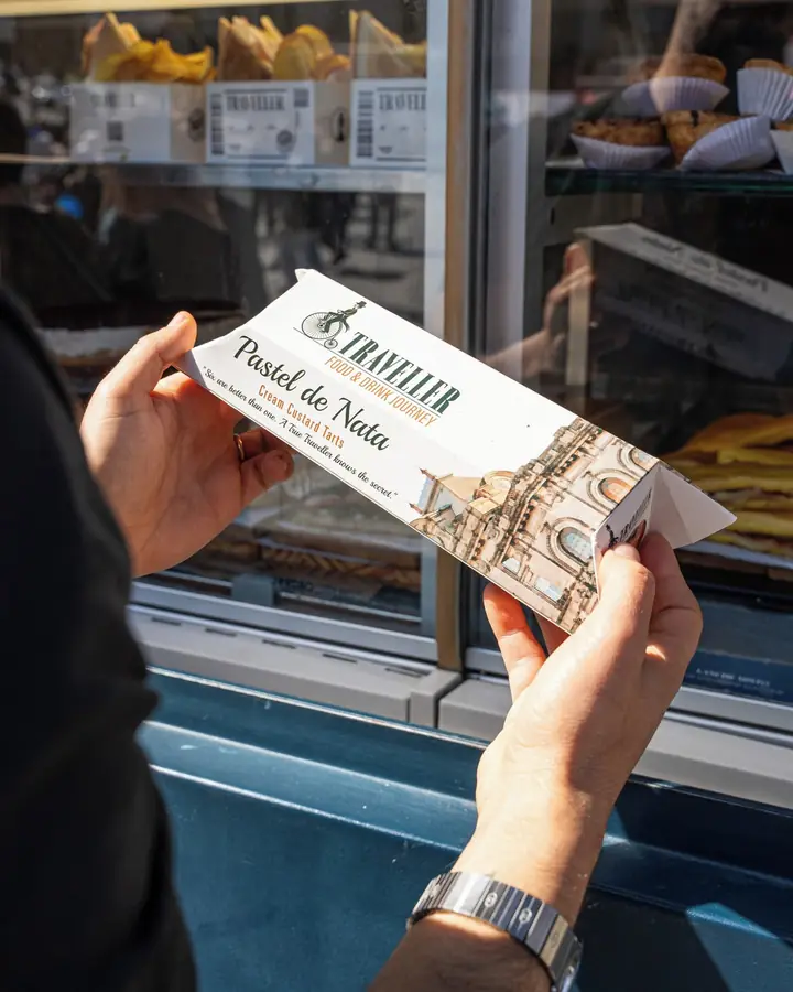 Hand holding a box of pastéis de nata to go, next to the window